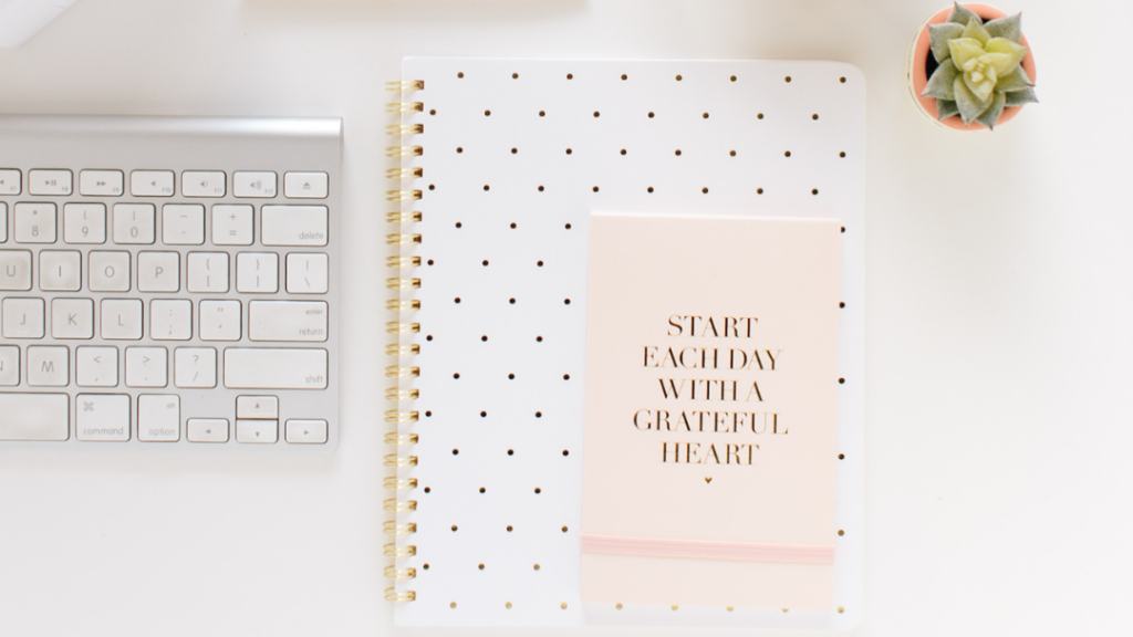 A wireless Apple keyboard with a notebook beside it that reads "Start each day with a grateful heart".