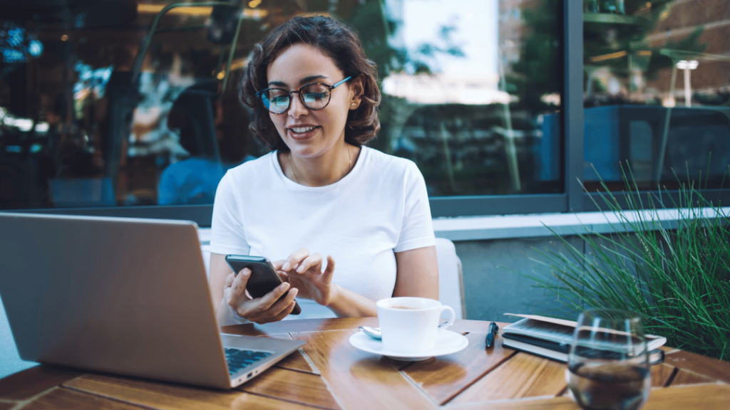 A woman wearing glasses sits at a table outside, focused on her phone, with a cup of coffee beside her, while she evaluates her digital notifications.