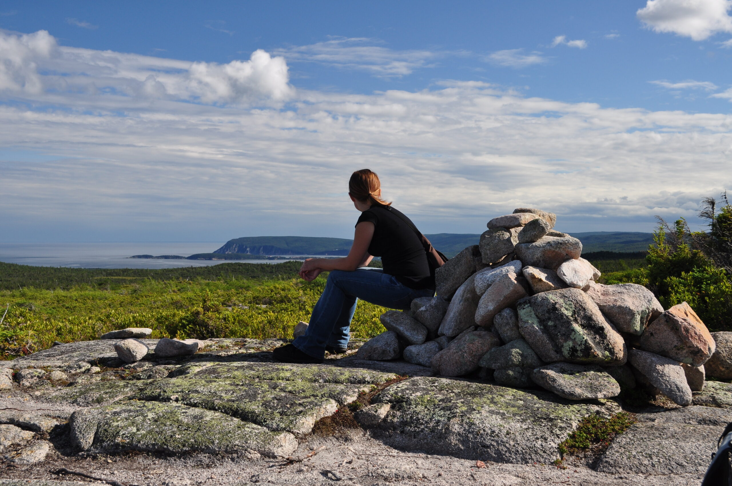 A serene image of a person sitting on a pile of rocks at the top of a mountain, gazing into the distance, symbolizing the peace and clarity that come from unplugging during a vacation. The breathtaking view highlights the beauty of nature and the importance of disconnecting from digital distractions to fully immerse oneself in the moment and recharge. Perfect for illustrating the concept of taking a proper vacation and finding balance in a tech-driven world.